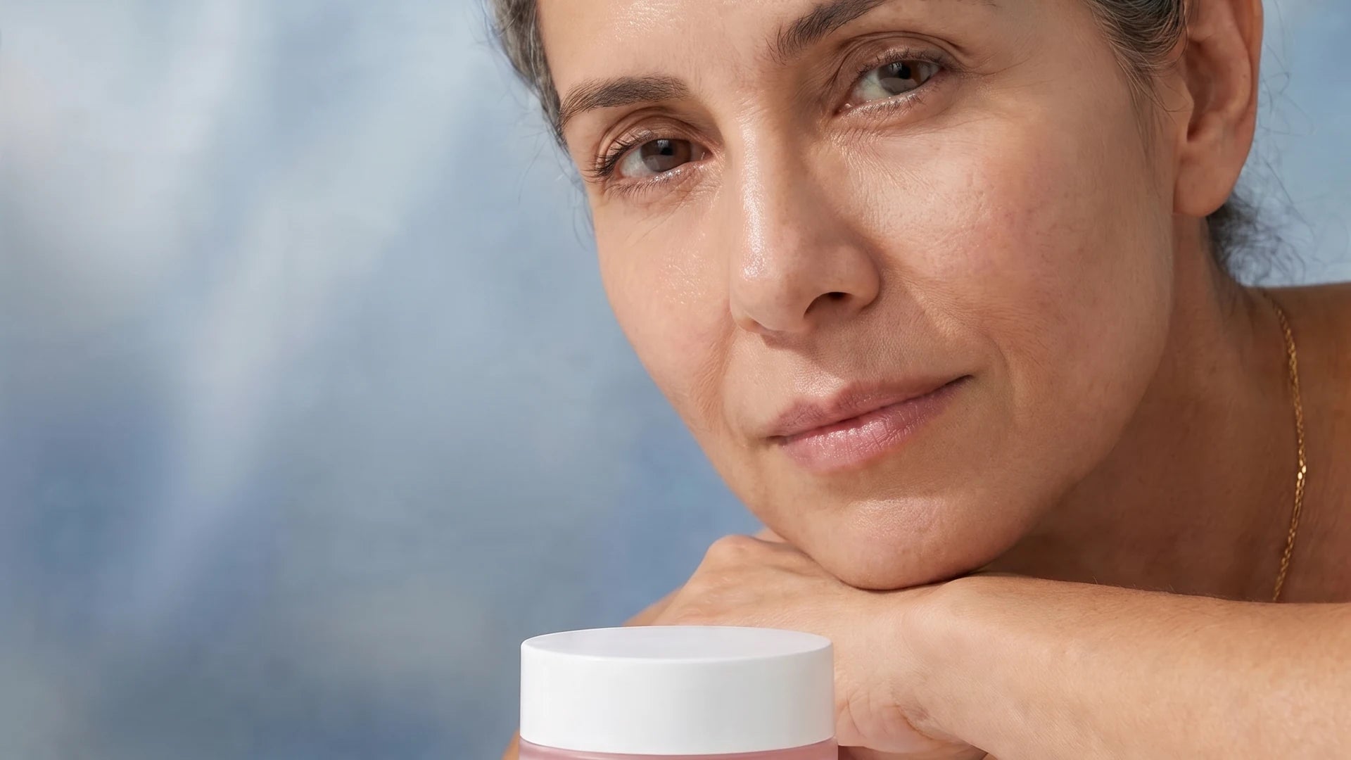 Mature woman with calm luminous skin resting chin on forearm beside SYVERA Bilayer Retinol Cream · cool editorial portrait · soft natural light